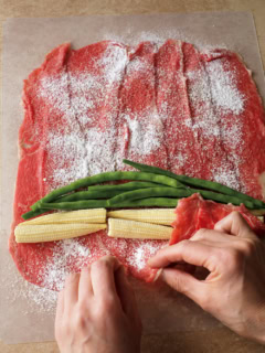 1 まな板に牛肉を1枚ずつ少し重なるようにして広げ、塩小さじ1/4、こしょう適量をふり、小麦粉適量を茶こしでふる。手前側にヤングコーンの根元と先端を互い違いに並べて置き、さやいんげんを牛肉の幅に揃えるようにして、左右に少し広げて置く。牛肉を手前から少しずつ持ち上げ、くるくると巻く。