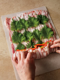2 豚肉はパックをそのままひっくり返し、まな板に広げる。塩小さじ1/4、こしょう適量をふり、小麦粉適量を茶こしでふる。青じそを全体に並べてのせ、手前側にパプリカを豚肉の幅に揃えるようにして、左右に少し広げて置く。豚肉を手前から少しずつ持ち上げ、くるくると巻く。